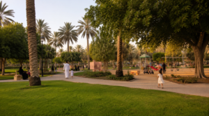 Al Nakheel Park in Riyadh with green lawns, palm trees, walking paths, playground swings and slides, and families enjoying the park in the evening.