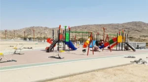 Public park playground with large multi-play towers, tube and spiral slides, swings and outdoor gym equipment on rubber safety flooring, with mountains behind.