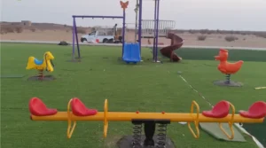 Yellow four-seat seesaw with red seats and two spring riders, a yellow horse and an orange duck, on artificial grass with a play tower and swing set behind.