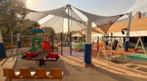 Shaded outdoor playground in Saudi Arabia with separate play areas for different ages, including toddler swings, spring riders, slides, climbing dome, monkey bars, and comfortable parent seating on safety flooring.