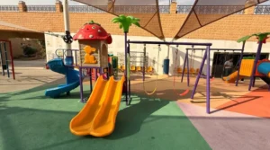Swing and slide set with iron frame, yellow double slide, blue spiral slide and strawberry-top play house, beside swings on colored rubber safety flooring under shade.