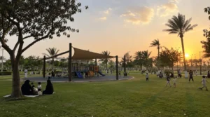 Tihama Park in Jeddah with shaded children’s playground, families sitting on grass, kids playing in an open green area, and sunset light.