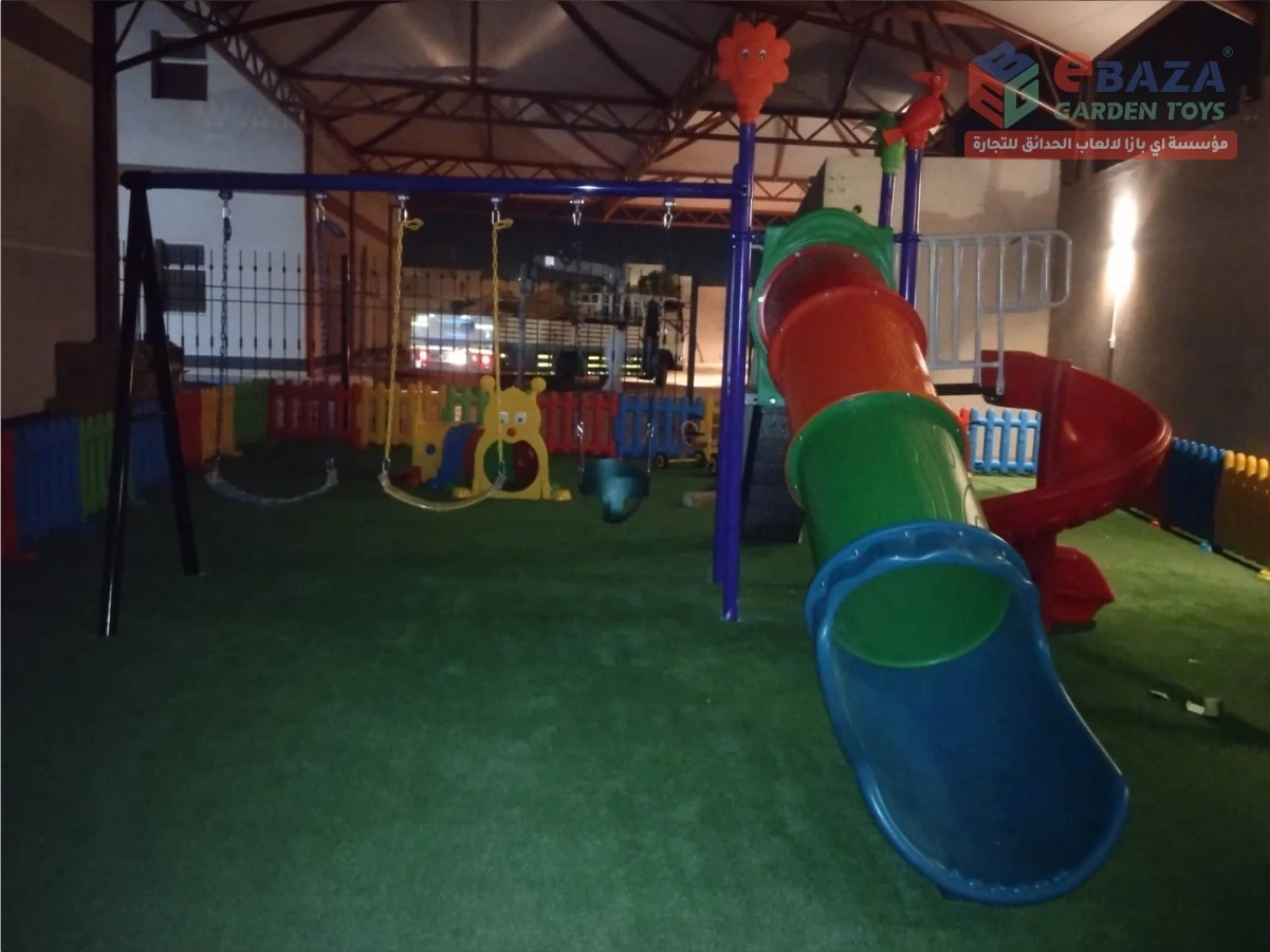 Indoor children’s playground with tube slide, spiral slide, swings, artificial grass flooring, and safety fence.