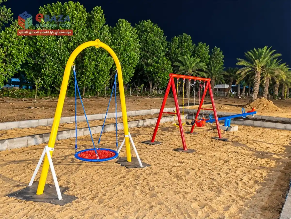 Wide view of a completed farmhouse playground with a yellow nest swing, red swing set, and blue seesaw on sand in Audat Sudair, Saudi Arabia