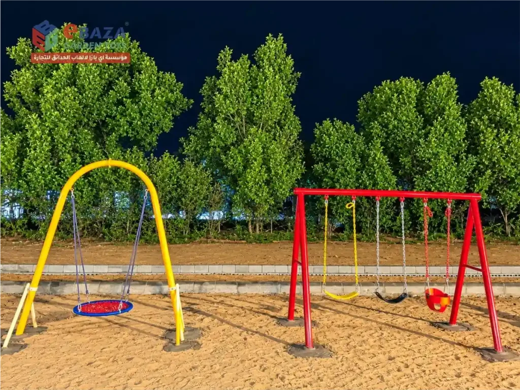 Red steel swing set with three seats and a blue rocking ride on a concrete block base in a sand playground at an Audat Sudair farmhouse